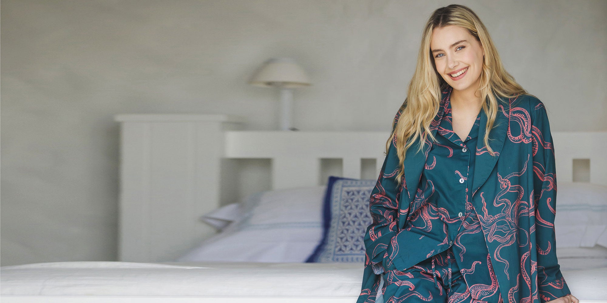 Woman in a patterned teal outfit sitting on a bed in a bedroom.