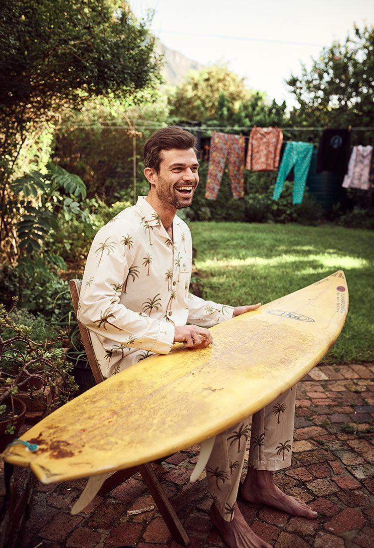 Man waxing his surfboard, wearing Palm Beach Long Pyjamas from Woodstock Laundry