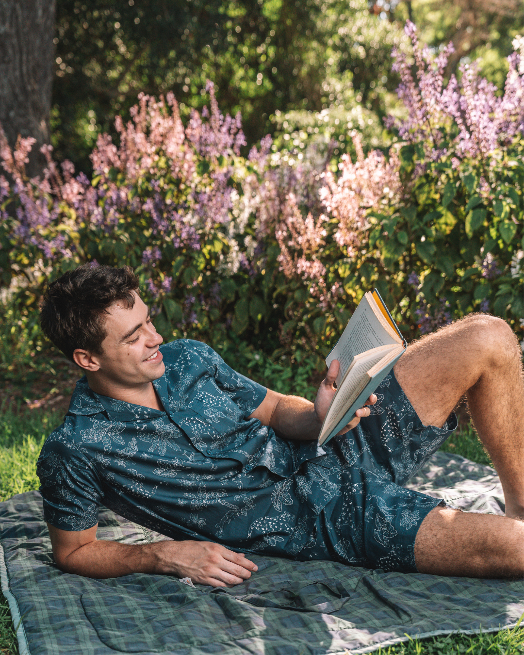 Man reading a book on a picnic blanket in a garden wearing a navy tropical leopard print organic cotton short pyjamas set